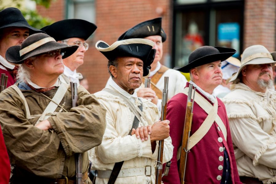 Line of individuals dressed in knee breeches, shirts, and tricorn hats typical to the 18th century play on the drums.