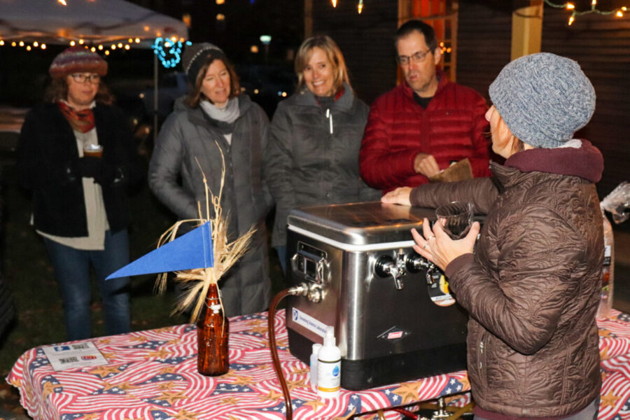 Brewer in a brown jacket stands behind a portable tap and pours for four Beer for History guests outside the Folsom Tavern