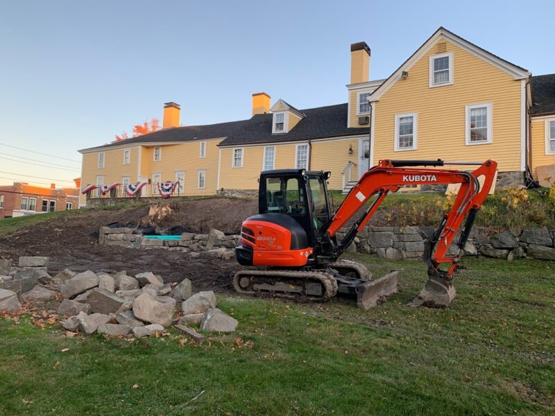 Large red construction vehicle sits on a grassy lawn with an embankment held in place by a stone wall of about 3 feet in height. On the hill above the ledge is the Ladd-Gilman House, a 300 year old building faces in cheery, yellow clapboard. 