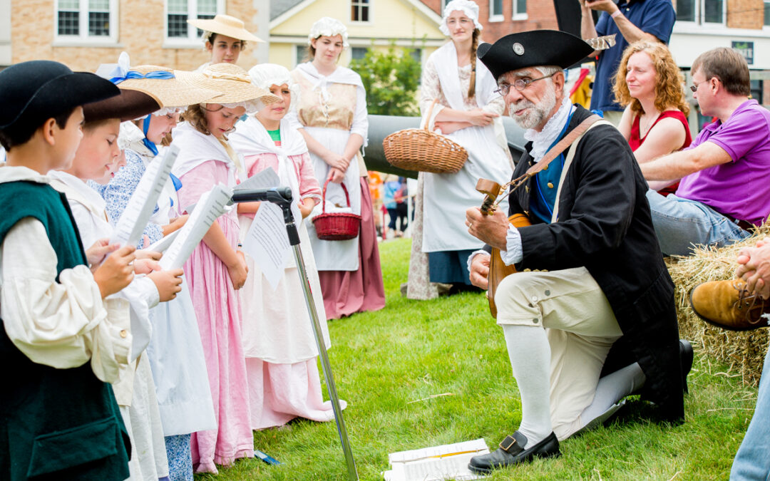 Man in 18th century clothing kneels on a lawn outside and palys a stringed instrument for a group of children.