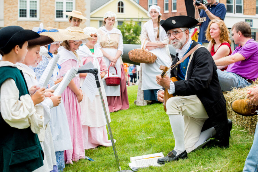 Man in 18th century clothing kneels on a lawn outside and palys a stringed instrument for a group of children.