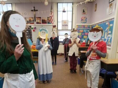 Elementary school aged children holding up paper plate masks with Revolutionary Era characters drawn on.