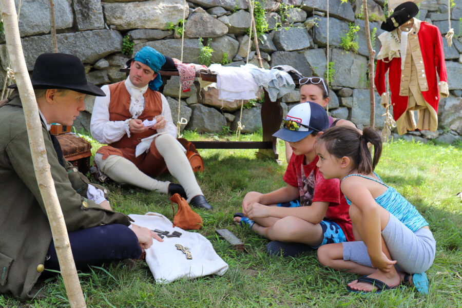 Two young children sit on the grass in front of two reenactors demonstrating 18th century tailoring