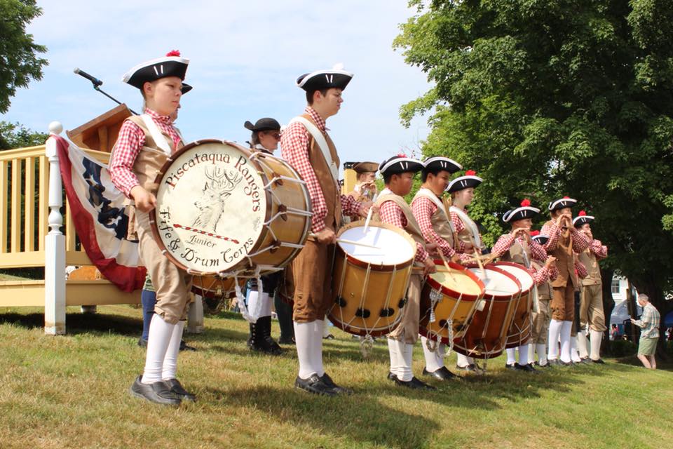 Line of individuals dressed in knee breeches, shirts, and tricorn hats typical to the 18th century play on the drums.