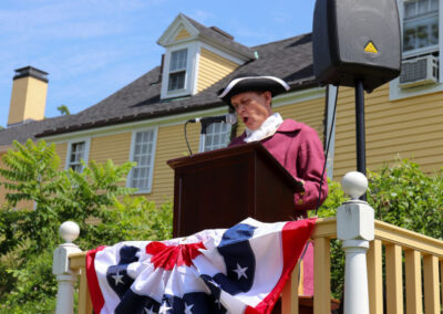 Person dressed in 18th century clothes as John Taylor Gilman stands at a podium with an American Flag draped in front of it reading from the Declaration of Independence on a sunny day.