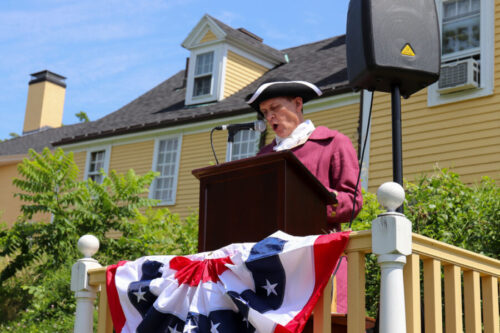 Person dressed in 18th century clothes as John Taylor Gilman stands at a podium with an American Flag draped in front of it reading from the Declaration of Independence on a sunny day.