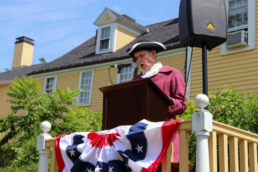 Person dressed in 18th century clothes as John Taylor Gilman stands at a podium with an American Flag draped in front of it reading from the Declaration of Independence on a sunny day.