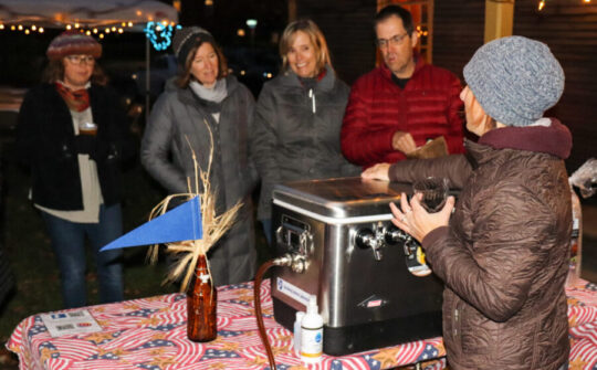 Brewer in a brown jacket stands behind a portable tap and pours for four Beer for History guests outside the Folsom Tavern