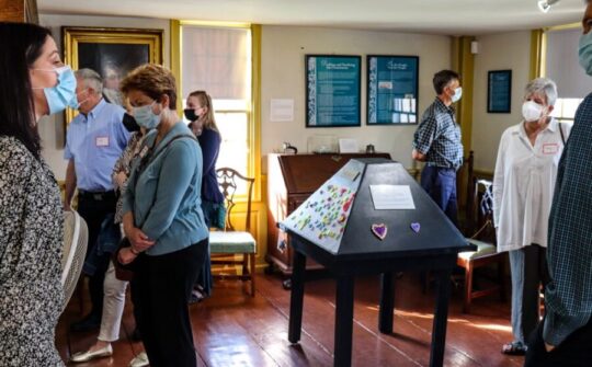 Seven individuals walk around a museum room with a pyramidal shaped interactive in the middle of the room