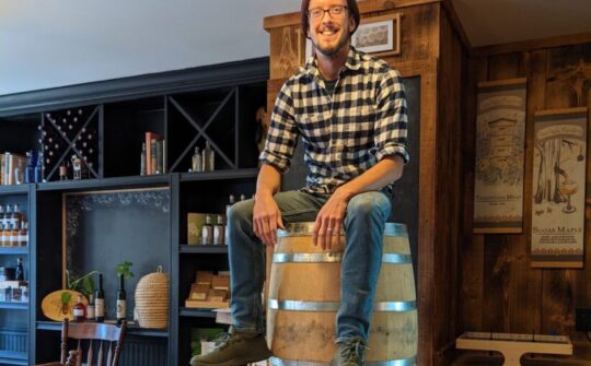 Matt Trahan of Sap House Meadery sits atop pyramidally stacked oak barrels wearing jeans, a flannel, and a grey beanie.