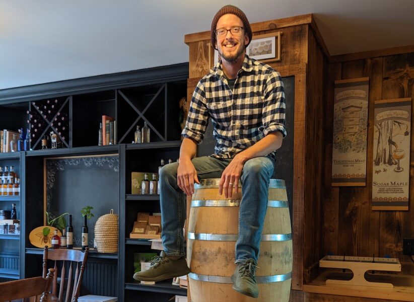 Matt Trahan of Sap House Meadery sits atop pyramidally stacked oak barrels wearing jeans, a flannel, and a grey beanie.