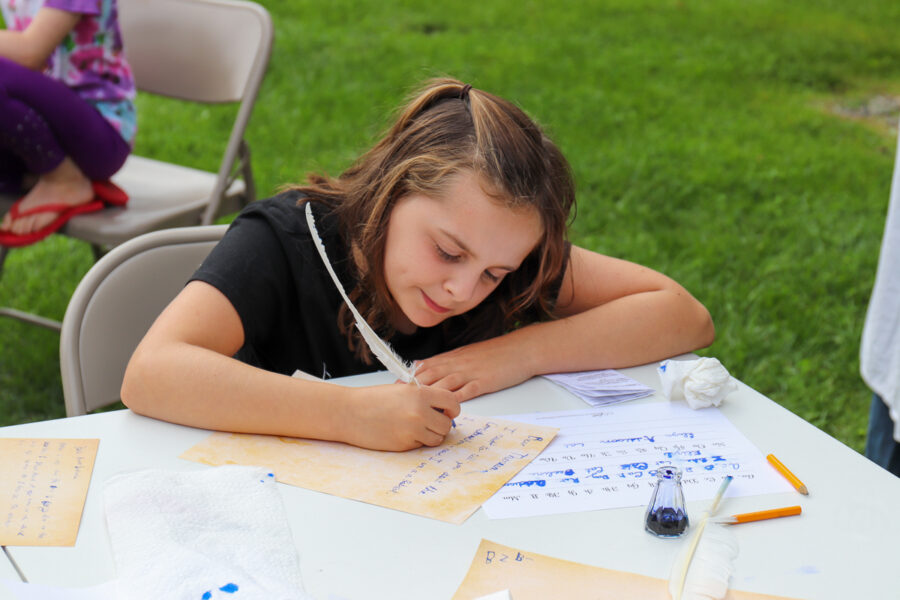 child sitting at a table outside, writing with a quill pen.